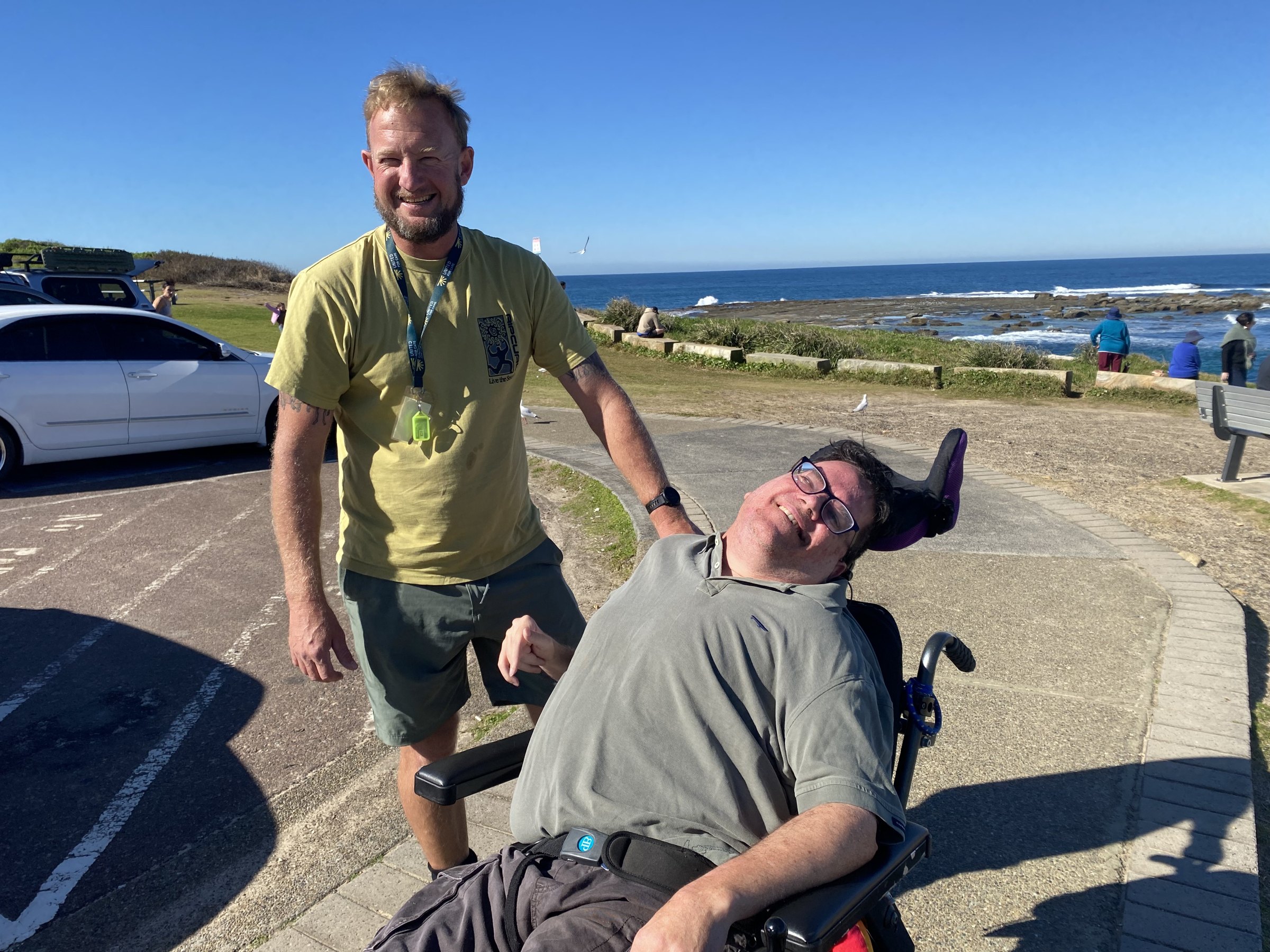 A support worker and a member smiling together on a coastal path by the ocean.